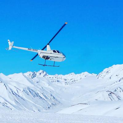 A white helicopter with 'N678AS' and 'ALASKA' written on its side flies over vast snow-covered mountains under a clear blue sky.