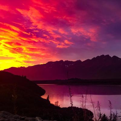 A breathtaking landscape photo captures a vivid sunset over a mountain lake. The sky is ablaze with bright yellow, orange, and magenta clouds, reflecting intensely on the calm water below. Dark silhouettes of mountains and hills frame the scene, with hints of snow on some distant peaks.