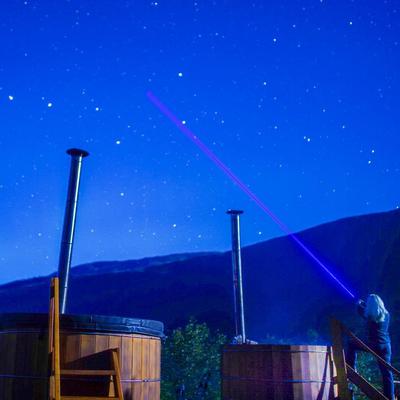 A person stands next to a wooden hot tub at night, pointing a bright purple laser beam into a clear, star-filled sky. Another hot tub with a chimney is visible to the left, with dark mountains in the background.