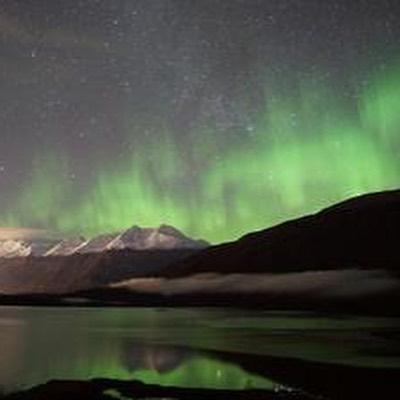 A nighttime landscape photo shows vibrant green aurora borealis streaking across a dark, star-filled sky above snow-capped mountains. The colorful lights and mountain peaks are reflected in the calm waters of a lake or river in the foreground.