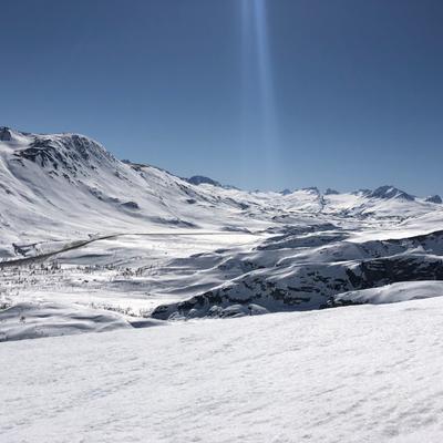 A panoramic view of the snow-covered Thompson Pass in Alaska, featuring towering mountains, a winding road through the valley, and a bright blue sky with a sun flare.