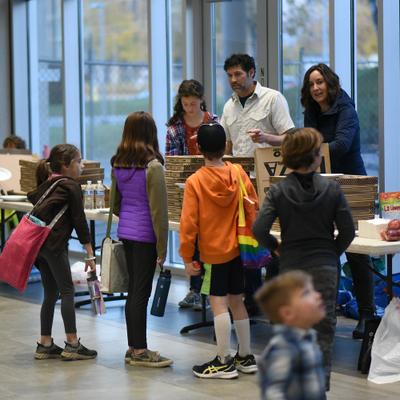 A diverse group of adults and children gather around tables with food and drinks at an indoor community event, likely during an Anchorage Assembly meeting. People are seen interacting and serving themselves from boxes and platters.