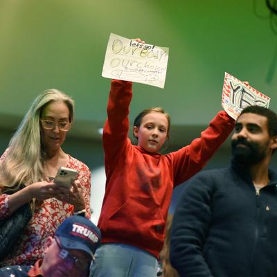 A young girl holds signs reading 'Our Body! OUR choice! I will not comply!' at an Anchorage Assembly meeting, surrounded by other attendees, including a woman looking at her phone and a man in a 'TRUMP' hat.