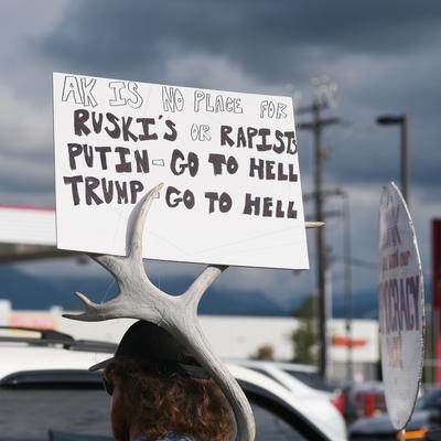 A person wearing a hat adorned with an antler holds a protest sign that reads 'AK IS NO PLACE FOR RUSKI'S OR RAPISTS PUTIN - GO TO HELL TRUMP - GO TO HELL' at a pro-Ukraine and anti-Trump rally in midtown Anchorage.