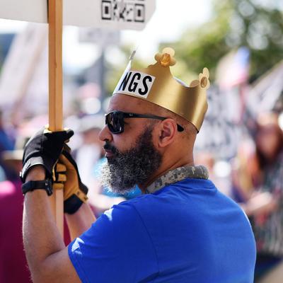 A man with a grey beard, sunglasses, and a gold paper crown (partially reading "NGS") holds a protest sign on a wooden stick. He is wearing a blue t-shirt and gloves, seen from the side at an outdoor community gathering in Anchorage.