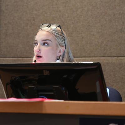 A young woman with blonde hair, glasses resting on her head, and a nose piercing looks intently to her left, partially obscured by a computer monitor on a desk in an indoor setting.