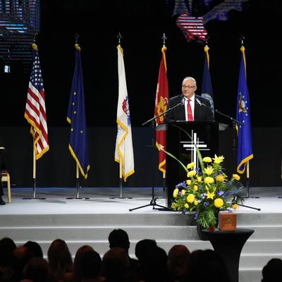 A man in a suit and glasses speaks at a podium adorned with a cross, flanked by the U.S. flag, Alaska state flag, and various military service flags. Other men are seated to the left of the speaker, and a vibrant yellow and blue floral arrangement is visible on the stage, with an audience partially visible in the foreground.