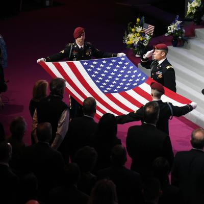 Military personnel in dress uniform present a large American flag during the memorial service for former U.S. Representative Don Young, with an urn on a stand and many attendees observing respectfully.