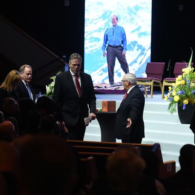 Attendees gather at a memorial service for Don Young, with an urn on a table, floral arrangements, and a large screen displaying a photo of Young against a mountain backdrop. American and Alaska flags are visible on the stage.