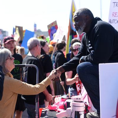 A diverse crowd gathers outdoors for the 'No Kings' protest in Anchorage, Alaska. A Black man on a platform leans down to shake hands with a white woman in the foreground, while other attendees and protest signs are visible in the sunny background.