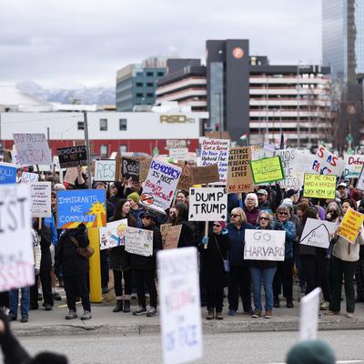 A large crowd of people gathers on a city street in Anchorage, Alaska, holding various protest signs. Distant snow-capped mountains and urban buildings are visible under an overcast sky.
