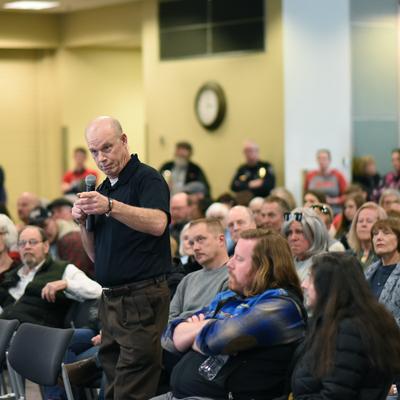 A man with a microphone addresses a diverse audience seated in rows at an indoor community town hall meeting in Anchorage.