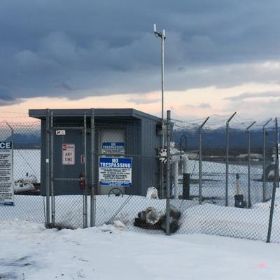 A snow-covered industrial facility, identified as Marathon Petroleum Corp at Anchorage Airport, is seen behind a chain-link fence with barbed wire. Distant snow-capped mountains are visible under a cloudy, twilight sky.