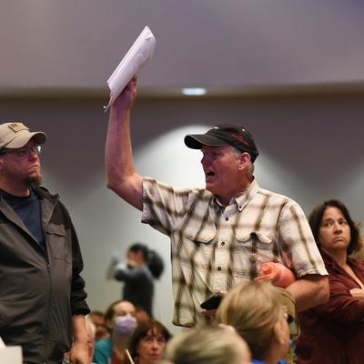 A man in a plaid shirt and baseball cap raises a document and speaks animatedly at an indoor public meeting, while other attendees listen and watch.