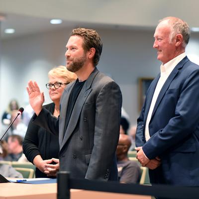 Three individuals stand at a podium in an assembly chamber, with one man in the center raising his right hand as if taking an oath. A woman stands behind him, and another man in a suit stands to his right, observing. An audience is visible in the background.