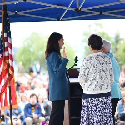 Suzanne LaFrance, wearing a teal blazer, stands at a podium with her right hand raised, being sworn in as mayor of Anchorage. To her right, two women face her, and in the background, a crowd of people watches under a blue canopy with flags, including the U.S. and Alaska flags, visible on the left.