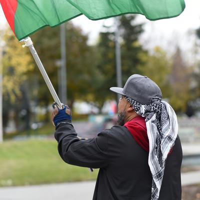 A person from behind, wearing a grey cap and keffiyeh, holds a red and green flag aloft during a protest in Anchorage. The background shows autumn trees and a park-like setting.