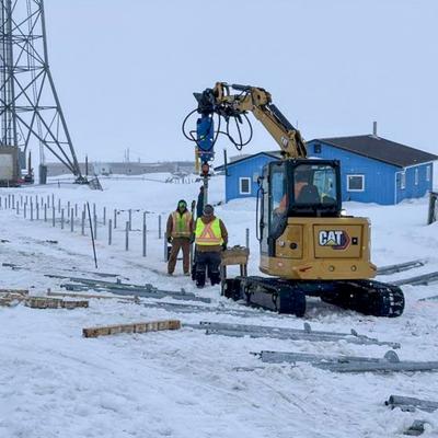 An excavator with a drilling attachment is used by construction workers to install metal posts in the snow-covered ground in Kipnuk, Alaska. Blue buildings and a large communication tower are visible in the background, indicating ongoing infrastructure development by the Alaska Department of Transportation & Public Facilities.