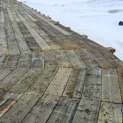 A long, weathered wooden boardwalk stretches into the distance over a snowy and icy landscape in Kwigillingok, Alaska. The planks are covered with some dirt and moss, and snow covers the ground on either side of the path.