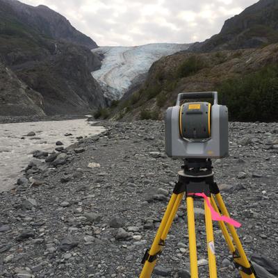 A gray and yellow surveying instrument on a tripod stands on a rocky riverbed in the foreground, with the vast, blue-white Exit Glacier visible in the background, nestled between rugged mountains under a cloudy sky.