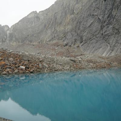 A vibrant turquoise glacial lake is nestled among steep, grey, rocky mountains under an overcast sky. The foreground features a rocky shore with a mix of grey and rust-colored boulders.