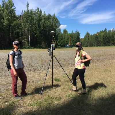 Two women in face masks and outdoor gear stand in a grassy field with a GPS surveying tripod between them, backed by a dense forest and a blue sky with clouds. An orange windsock is visible in the distance, suggesting an airfield.