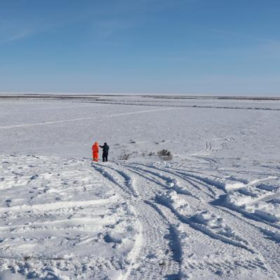 Two individuals stand on a snow-covered bank, observing a vast, frozen landscape under a clear blue sky. Vehicle tracks lead down towards the distant Colville River, suggesting preparations for winter access.