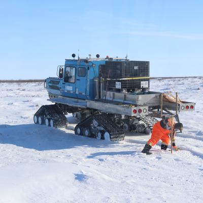 A person in bright orange winter gear kneels in a vast, snow-covered landscape, carefully placing a node into the ground. Behind them, a large blue tracked vehicle, equipped with various supplies, is parked under a clear blue sky.