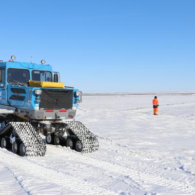A blue Tucker Sno-Cat, a large tracked vehicle, is parked on a vast, snow-covered plain under a clear blue sky. A person in orange high-visibility gear stands in the distance, surveying the expansive winter landscape of the Colville River bluff in Arctic Alaska.