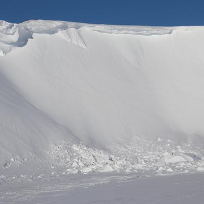 A towering, snow-covered bluff along the Colville River dominates the frame under a clear blue sky. A large pile of snow and ice debris, likely from an avalanche or cornice collapse, lies at the base of the steep slope.